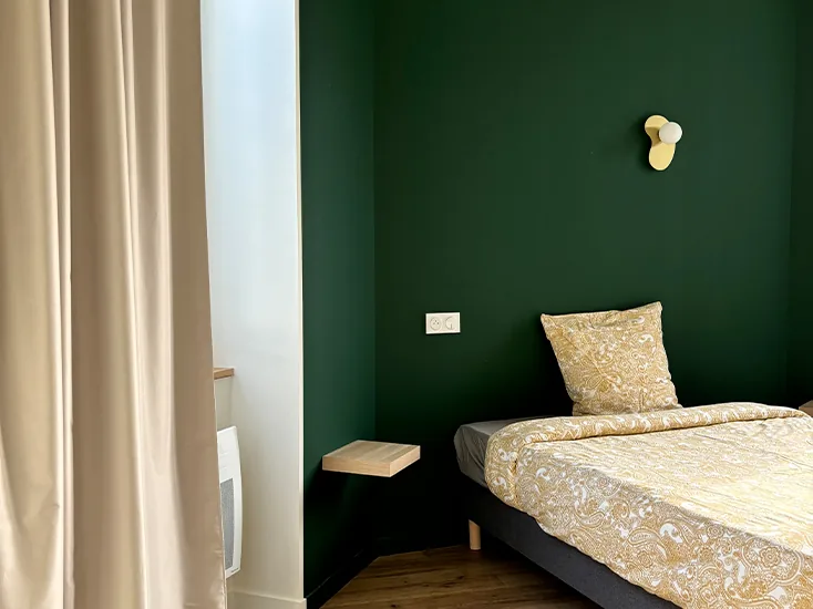 coliving-chambre-verte-fenetre-le-chateau-bayonne Chambre au mur vert avec lit simple, applique murale et rideau clair